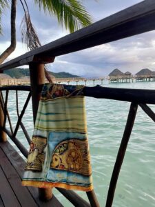 Maison Devienne silk scarf over a wooden railing, facing a Polynesian lagoon with overwater bungalows in the background.